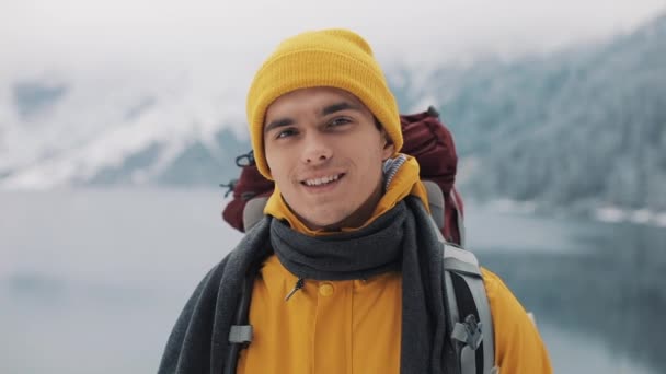 Porträtt av en ung attraktiv man i vinterkläder. Vandring man klädd i gul vinterkläder ser på kameran och leenden. Vackra berg och snötäckta sjön bakgrund — Stockvideo
