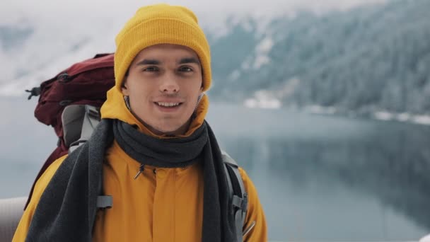 Potret seorang pemuda menarik dalam pakaian musim dingin. Pria pendaki yang mengenakan pakaian musim dingin kuning melihat ke kamera dan tersenyum. Indah gunung dan salju-tertutup danau latar belakang — Stok Video