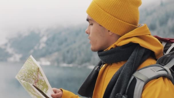 Viajando por las montañas. Joven excursionista utiliza un mapa en papel de la zona. Busca en el camino. Disfruta de la aventura y los viajes. Sobre un fondo de montañas de invierno y un hermoso lago — Vídeos de Stock