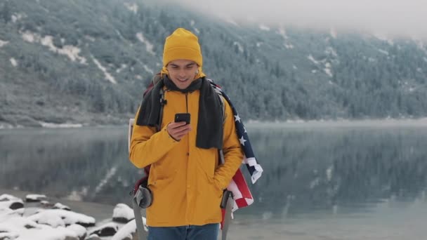 ハイキング冬ツアーでスマート フォンを使用してアメリカの旅行者の男。雪に覆われた湖と背景の山です。旅行・通信の概念: ハイカーのスクロールと携帯電話で行をタップ — ストック動画