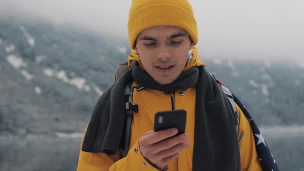 Gelukkig reiziger man met smartphone in winter wandeltocht. Besneeuwde wazig bergen cape en meer. Reizen en communicatie concept: wandelaar schuiven en op de lijn te tikken door cell phone — Stockvideo