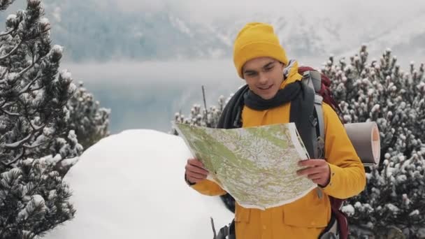 Viajar nas montanhas. Jovem vestindo roupas amarelas de inverno segurando mapa em pé na floresta coberta de neve. Viagem, aventura, escalada — Vídeo de Stock