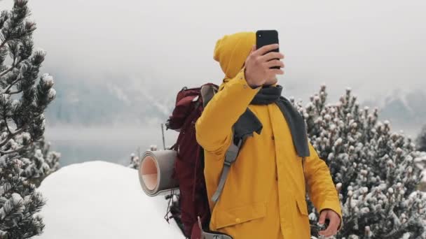 Красивий чоловік-поводир відео дзвінки на смартфон, махаючи на камеру, посміхаючись і розмовляючи з друзями. Прекрасні зимові гори і озеро на фоні, повільний рух — стокове відео
