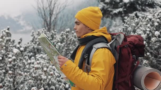 山の旅。森の中に立っている地図を保持している黄色の冬の服を着ている若い男は雪で覆われています。旅行、登山、冒険 — ストック動画