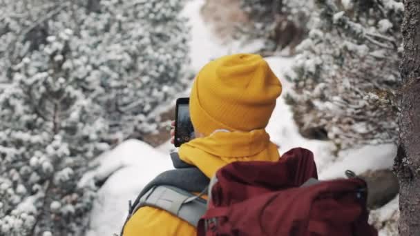 在山里旅行。身穿黄色冬装的年轻人走在被雪覆盖的智能手机上拍摄视频的树林里。旅行, 冒险, 攀岩。后视图 — 图库视频影像