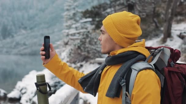 Молодой привлекательный мужчина в желтой куртке стоит в красивых горах. Турист восхищается природой и делает фотографии — стоковое видео