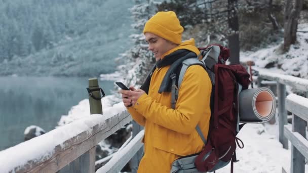 Homem viajante jovem usando smartphone em caminhadas passeio de inverno. Montanhas nevadas nevadas capa e lago. Conceito de viagem e comunicação: caminhante rolagem e tocando on-line por telefone celular — Vídeo de Stock