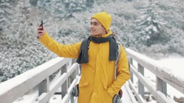 Yakışıklı uzun yürüyüşe çıkan kimse adam video smartphone, kamera, gülümseyen ve arkadaşlarınızla konuşma sallayarak çağırma. O köprüde duruyor. Güzel kış dağlar bir arka plana dayanır. Ağır çekim