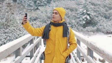Yakışıklı uzun yürüyüşe çıkan kimse adam video smartphone, kamera, gülümseyen ve arkadaşlarınızla konuşma sallayarak çağırma. O köprüde duruyor. Güzel kış dağlar bir arka plana dayanır. Ağır çekim