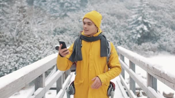 ハンサムなハイカー男のビデオ通話スマート フォン、カメラ、笑みを浮かべて、友人同士の会話で手を振るします。彼は橋の上に立っています。背景の美しい冬の山。スローモーション — ストック動画