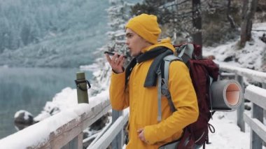 Onun elinde bir akıllı telefon ile karlı dağlarda bir köprüde duran genç bir turist. Konuşmayı ile sesli mesaj yazmak dışında Smartphone fiyatı