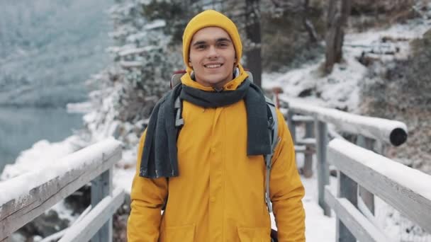 Портрет молодого привлекательного мужчины в зимней одежде. Турист в жёлтой зимней одежде смотрит в камеру и улыбается. Он стоит на мосту. . — стоковое видео