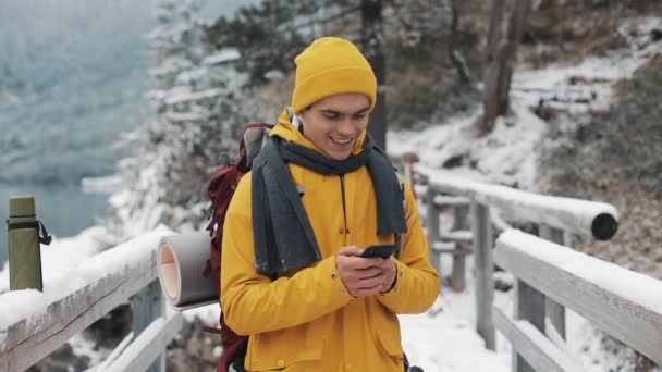 Egy fiatal turista, egy híd, a havas hegyek és használ smartphone. Gyönyörű hegyek, háttér — Stock videók