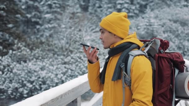 A young tourist standing on a bridge in the snowy mountains with a smartphone in his hands. Hiker having conversation with voice message recorder on smartphone outside — Stock Video