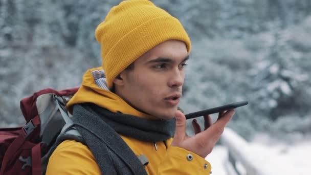 Un jeune touriste debout sur un pont dans les montagnes enneigées avec un smartphone à la main. Randonneur ayant une conversation avec un enregistreur de messages vocaux sur smartphone à l'extérieur — Video