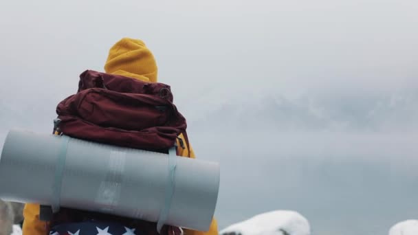 在山里旅行。身穿黄色冬衣的年轻人在山湖附近的石头上行走。冬季旅行, 冒险, 攀岩。后视图 — 图库视频影像