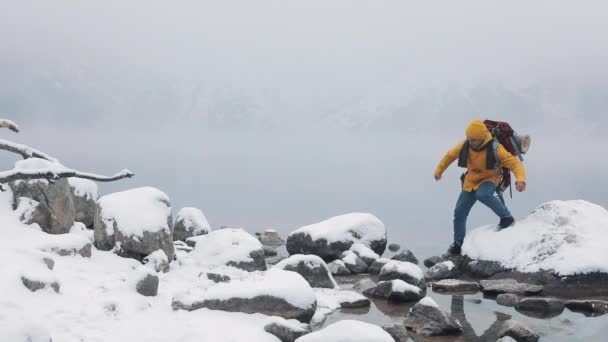 Voyager dans les montagnes. Jeune homme portant des vêtements d'hiver jaunes marchant sur des pierres près du lac de montagne. Temps d'hiver Voyage, aventure, escalade — Video