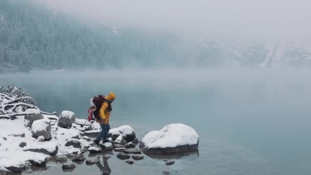 Американские туристы в жёлтой зимней одежде ходят по камням возле горного озера и поднимают руки на концепцию победителя. Пеший поход зимнего альпиниста — стоковое видео