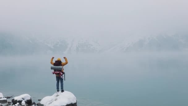 American hiker stands on a stone near a mountain lake and raising hands on mountain top winner concept. Snowy blurred mountains cape and lake — Stock Video