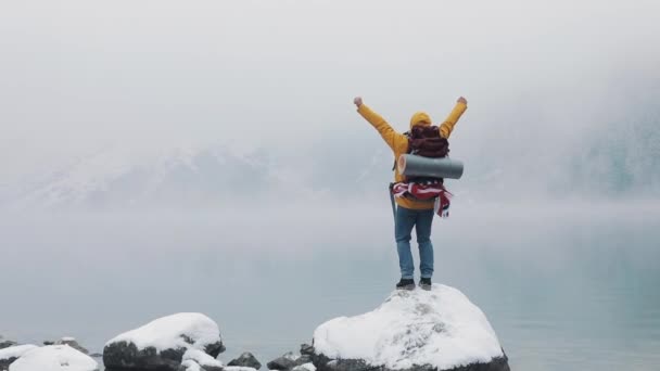 American hiker stands on a stone near a mountain lake and raising hands on mountain top winner concept. Snowy blurred mountains cape and lake — Stock Video