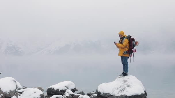 Hombre viajero feliz usando teléfono inteligente en la excursión de invierno senderismo. Caminante americano se encuentra en una piedra cerca de un lago de montaña. Montañas nevadas borrosas capa y lago. Concepto de viaje y comunicación — Vídeo de stock