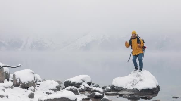 Video verticale. Bell'uomo americano escursionista in piedi sulla pietra vicino al bellissimo lago e divertenti balli. Belle montagne invernali sullo sfondo. Rallentatore — Video Stock