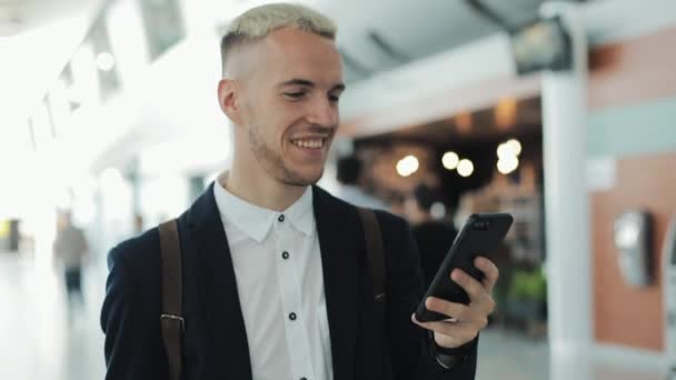 愉快的商人在机场等候登机时使用智能手机。游客经过机场的航站楼 — 图库视频影像