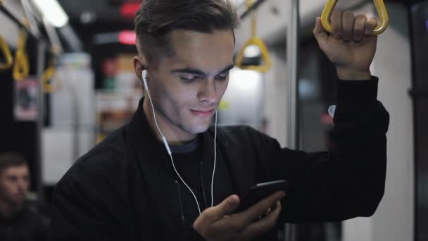 Retrato de hombre atractivo en auriculares sostiene la barandilla, escuchando música y navegando en el teléfono móvil en el transporte público. Ciudad luces fondo — Vídeo de stock