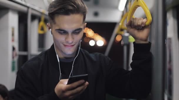 戴耳机的迷人男子的肖像拿着扶手, 听音乐, 在公共交通中用手机浏览手机。城市灯光背景 — 图库视频影像