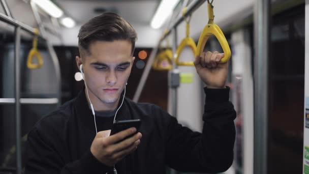 Retrato de un hombre serio con auriculares sostiene la barandilla, escuchando música y navegando en el teléfono móvil en el transporte público. Ciudad luces fondo — Vídeo de stock