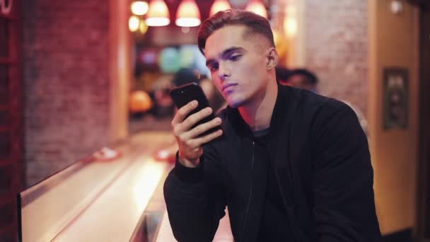 Triste jovem atraente homem usando Smartphone na noite. Ele está sentado em um bar ou restaurante perto de sinalização de néon. Fechar — Vídeo de Stock