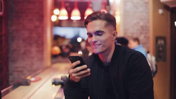 Young Attractive Man with Beautiful Smile is Using Smartphone at Evening. He sitting in a bar or restaurant near neon signage. Close up — Stock Video