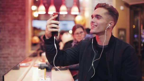 Beau jeune homme utilise un casque et son smartphone pour un appel vidéo. Il est assis au bar près des enseignes au néon. Ambiance joyeuse, communication moderne, technologies, appareils. Portrait masculin — Video