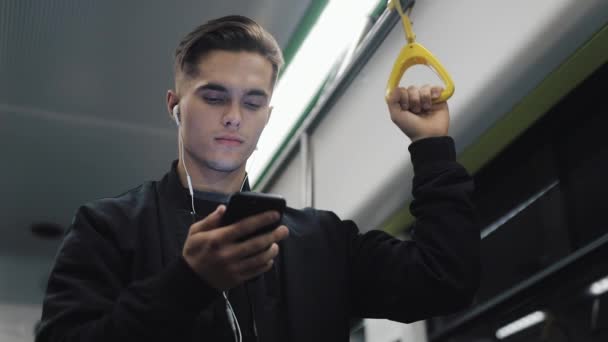 Portrait de l'homme sérieux dans les écouteurs tient la main courante, écouter de la musique et la navigation sur le téléphone mobile dans les transports publics. Fond de lumières de ville — Video