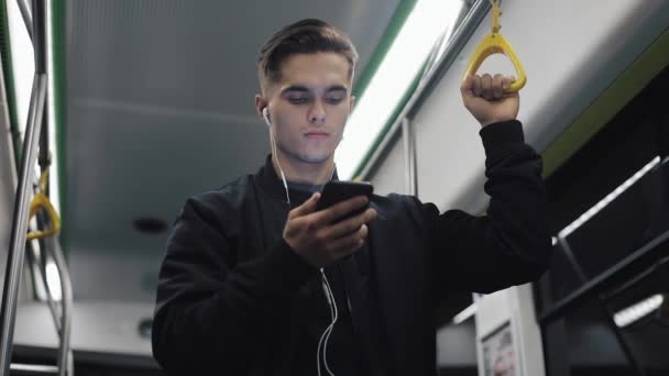 Kulaklığımda ciddi adam portresi Küpeşte, müzik ve hareket eden telefon içinde toplu taşıma üzerinde tarama tutar. Şehir ışıkları arka plan — Stok video