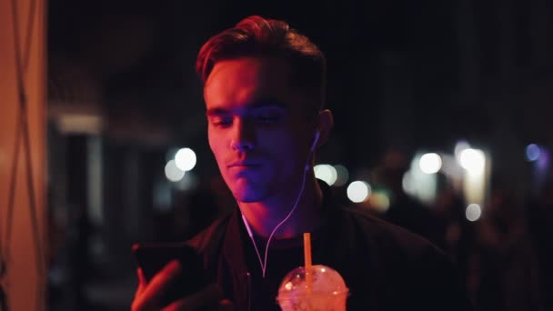 Przystojny, młody człowiek z słuchawki za pomocą smartfona dla rozmów wideo z przyjaciółmi. On stoi wieczorem na ulicy w pobliżu neony — Wideo stockowe