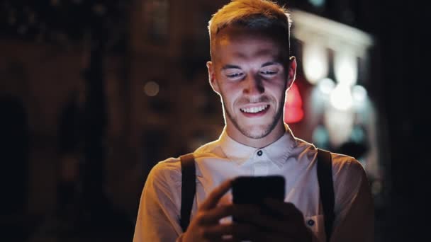夜の街の通りに立っているスマート フォンを使用しての若い魅力的な男。コミュニケーション、旅行、観光、デート、ビジネス コンセプト — ストック動画