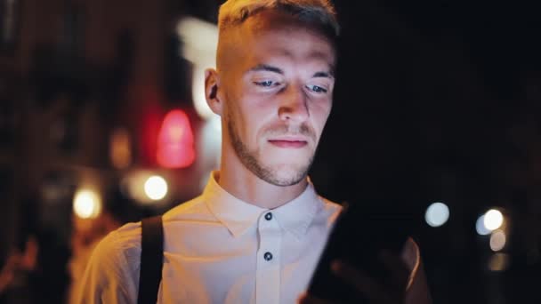 Jovem homem atraente usando smartphone andando na rua da cidade noturna. Comunicação, viagens, turismo, namoro, conceito de negócio — Vídeo de Stock