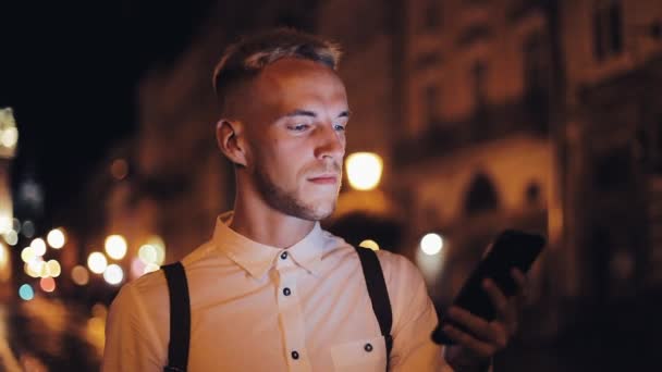 夜の街の通りの上を歩いてのスマート フォンを使用しての若い魅力的な男。コミュニケーション、旅行、観光、デート、ビジネス コンセプト — ストック動画