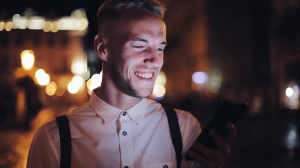 Joven hombre atractivo usando teléfono inteligente de pie en la calle de la ciudad nocturna. Comunicación, viajes, turismo, citas, concepto de negocio — Vídeos de Stock