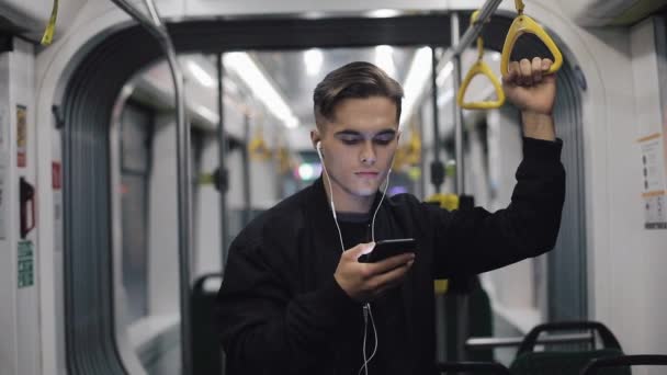 Retrato de homens bonitos em fones de ouvido ouvindo música e navegando no celular em transportes públicos. Ele segura o corrimão e dança engraçada — Vídeo de Stock