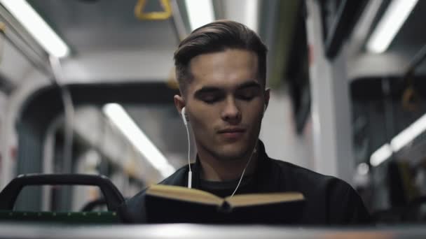 Joven hombre guapo sentado en el transporte público leyendo un libro - viajero, estudiante, concepto de conocimiento. Joven con auriculares en el tranvía leyendo un libro — Vídeo de stock