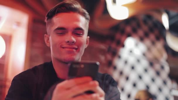 Aantrekkelijke jongeman met behulp van Smartphone op de avond. Hij zit in een bar of restaurant in de buurt van neon bewegwijzering. Close-up — Stockvideo