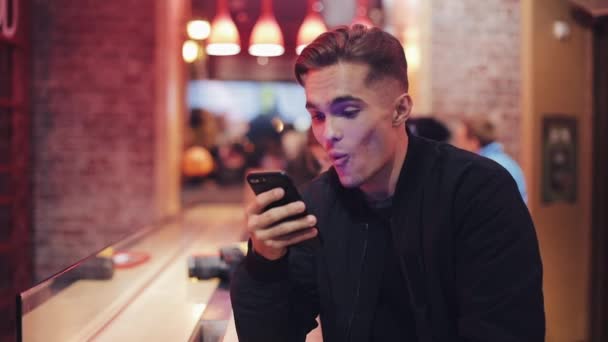 Joven hombre atractivo con hermosa sonrisa está utilizando el teléfono inteligente en la noche sentado en un bar o restaurante cerca de la señalización de neón. Está leyendo buenas noticias. De cerca. — Vídeo de stock