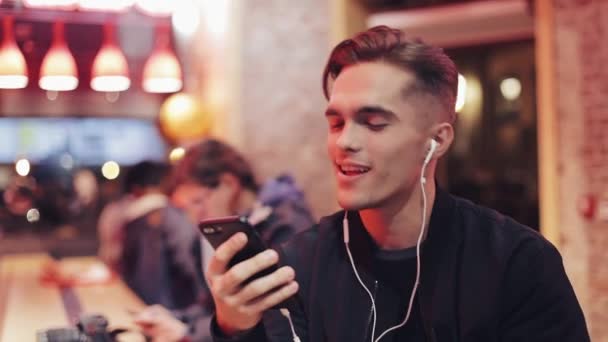 Beau jeune homme utilise un casque et son smartphone pour un appel vidéo. Il s'assoit au bar près des enseignes au néon et boit du coctail. Ambiance joyeuse, communication moderne, technologies, appareils — Video