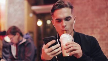 Yakışıklı genç kulaklık ve onun smartphone için video çağrısı kullanır. Neon işaret ve içki kokteyl barda oturacak. Neşeli ruh, modern iletişim, teknolojiler, cihazlar