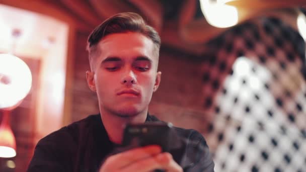 Серьезный молодой привлекательный человек, использующий смартфон на вечере. Он сидит в баре или ресторане рядом с неоновыми вывесками. Закрыть — стоковое видео