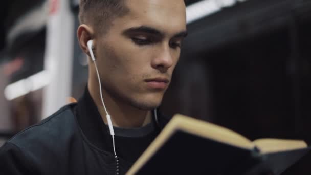 Joven hombre guapo sentado en el transporte público leyendo un libro - viajero, estudiante, concepto de conocimiento. Joven con auriculares en el tranvía leyendo un libro — Vídeos de Stock