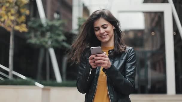 Joven mujer feliz caminando en la calle de la ciudad mientras usa el teléfono inteligente. Movimiento lento — Vídeos de Stock