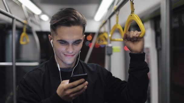 Portrait of attractive man in headphones holds the handrail, listening to music and browsing on mobile phone in public transport. City lights background — Stock Video